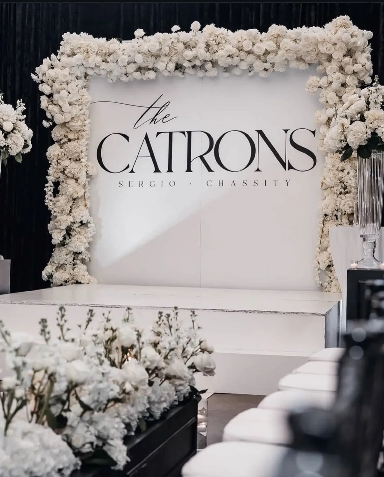 A white wedding welcome backdrop reading 'The Catrons' framed by white florals
