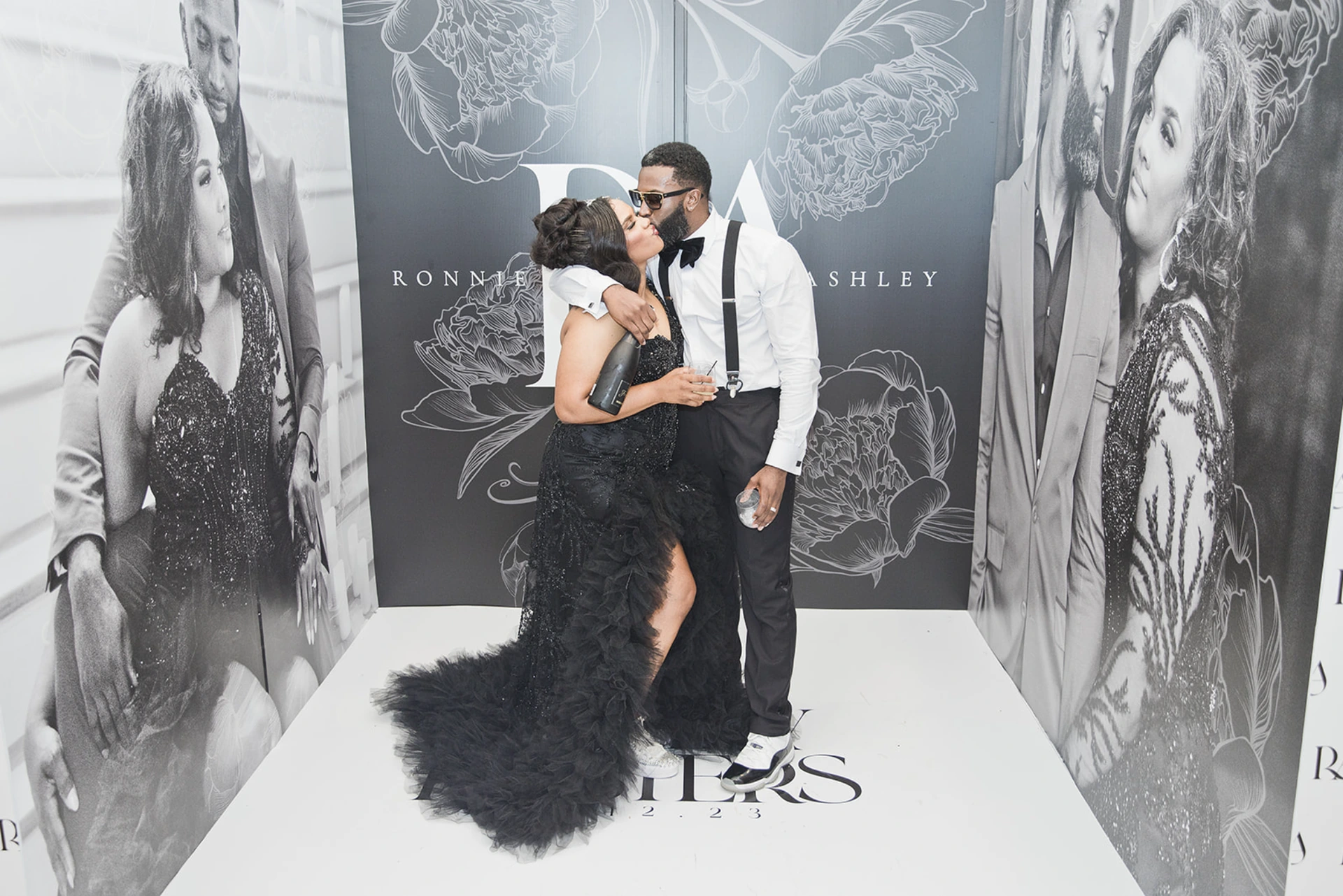 A couple embracing in front of a custom step-and-repeat backdrop printed with their monogram and portraits