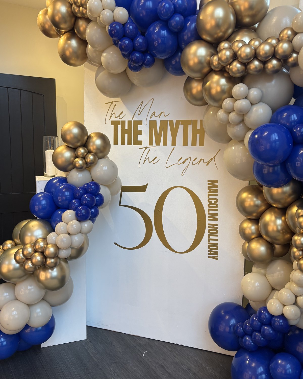 A white 50th birthday backdrop reading The Man, The Myth, The Legend, framed by a blue and gold balloon garland
