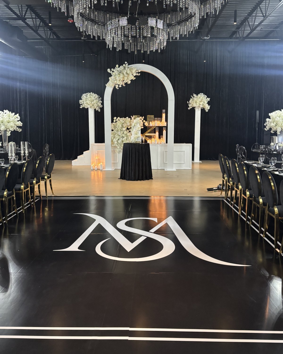 A black dance floor wrap with a white monogram in front of a floral arch at a wedding reception